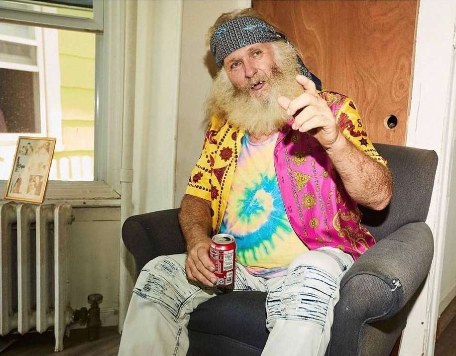 Elderly man with a long white beard sits in a gray armchair holding a Coca-Cola can wearing tie-dye and a bandana indoors.
