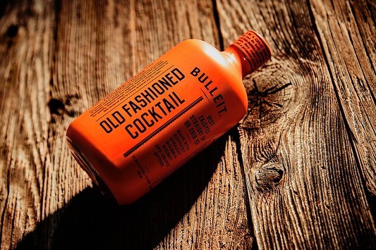 Bulleit Old Fashioned Cocktail bottle on a rustic wood surface. Close-up of the bright orange bottle with dark brown lettering.