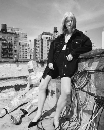 Model standing on a city rooftop wearing a dark denim jacket and shorts, with a second person seated wearing reflective silver attire
