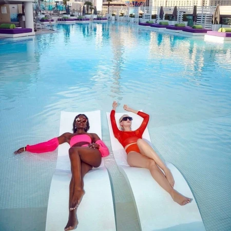 Two women relaxing on white loungers submerged in a sparkling rooftop swimming pool with purple sunbeds