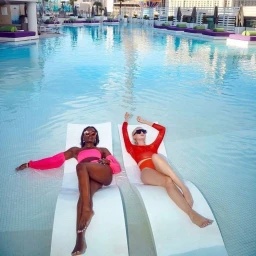 Two women relaxing on white loungers submerged in a sparkling rooftop swimming pool with purple sunbeds