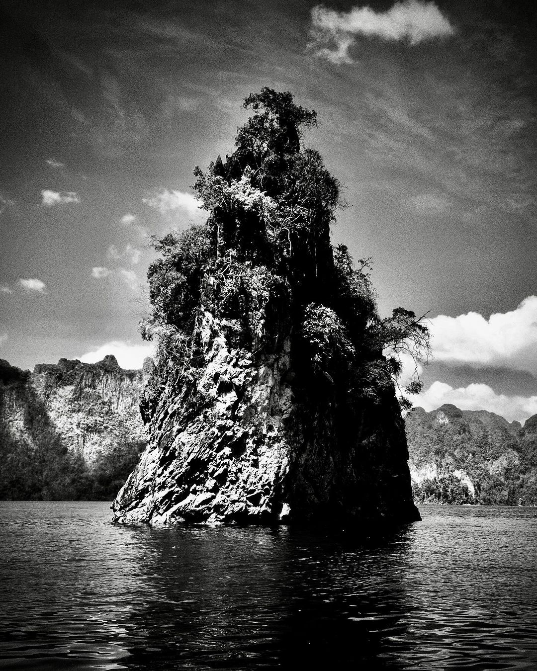 Monochromatic photo of a lush, rocky island in a lake, set against a backdrop of rolling hills and a sky filled with scattered clouds.