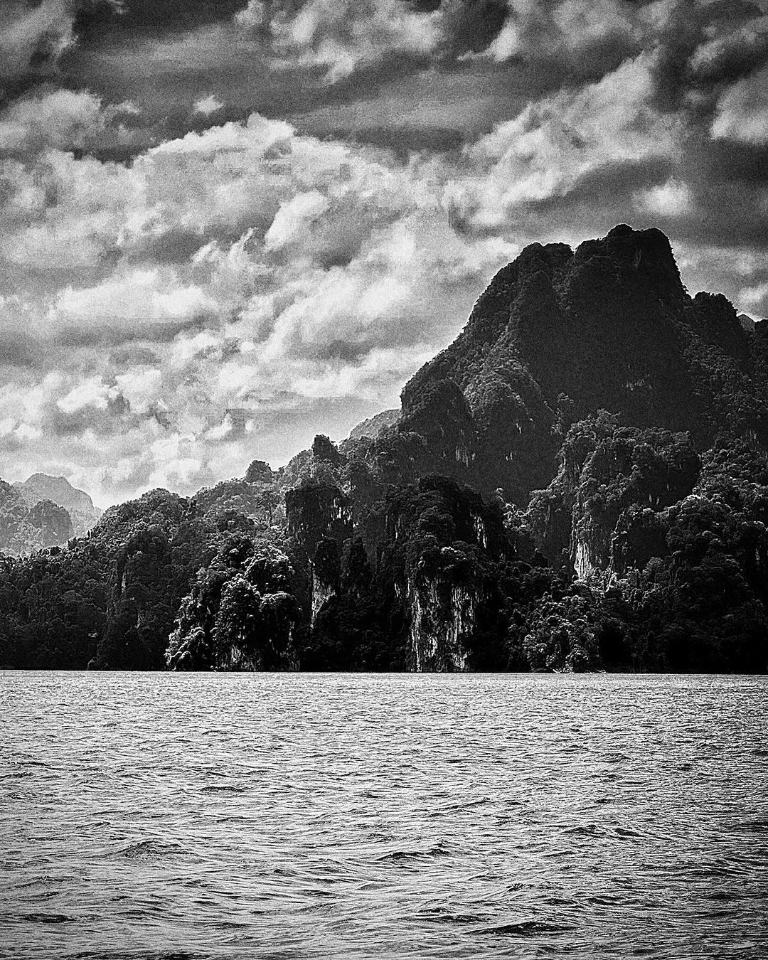Black and white shot of a mountain range covered in lush vegetation, rising from a body of water under a sky full of clouds.