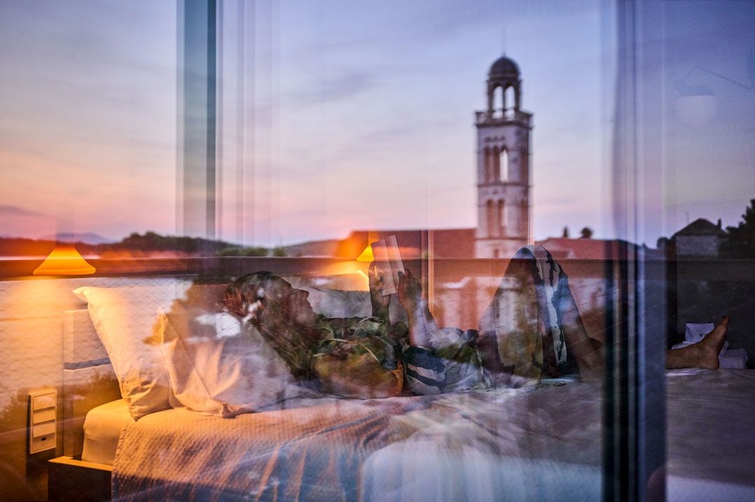 A woman lies on a bed in a sunlit room with a distant tower visible through the window, creating a dreamy and serene atmosphere.