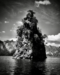 Monochrome vertical photograph of a dramatic rocky karst formation covered in vegetation rising out of dark water under a cloudy sky.