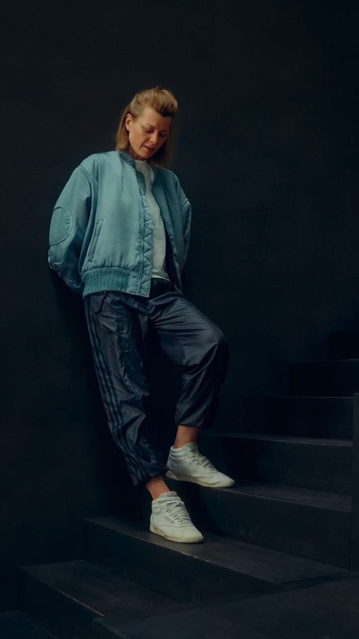 Woman in a blue jacket and dark track pants, leaning on a dark staircase in white sneakers, looking down in a fashion-style shot.