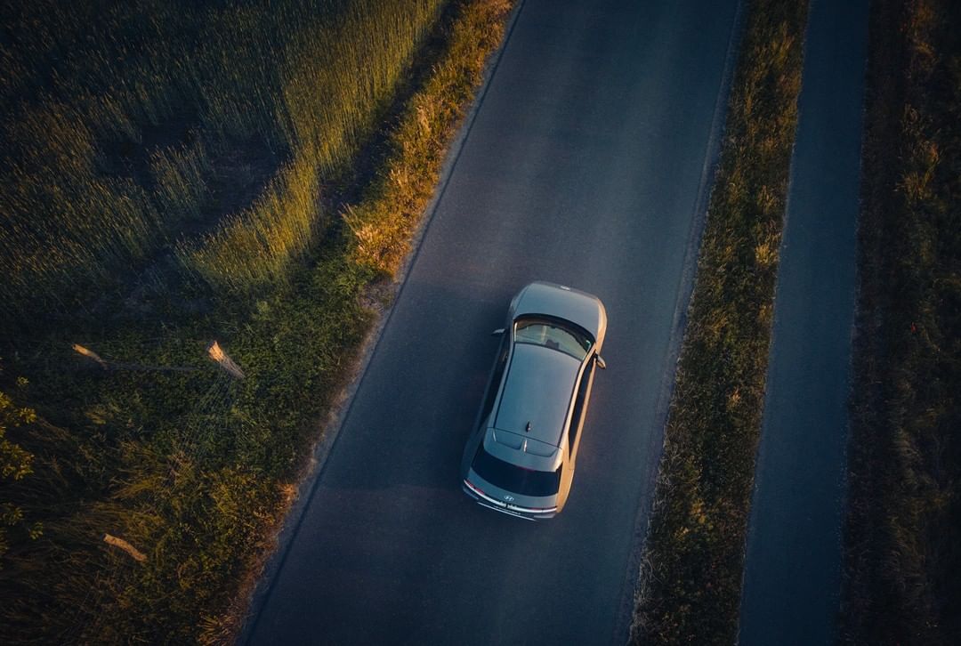 Aerial shot of a silver car on an asphalt road, flanked by green fields with golden sunlight edges during late afternoon.