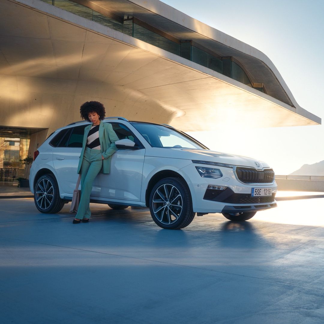 A white Skoda SUV parked outdoors with an adult woman in a light green suit leaning on the hood in front of a modern building.