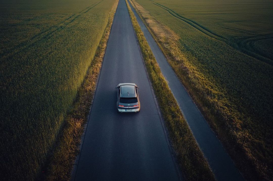 Overhead aerial shot of a gray car driving on a rural highway through green fields under soft, diffused light at dusk or dawn.