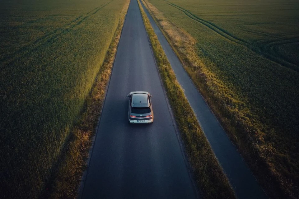 Overhead aerial shot of a gray car driving on a rural highway through green fields under soft, diffused light at dusk or dawn.