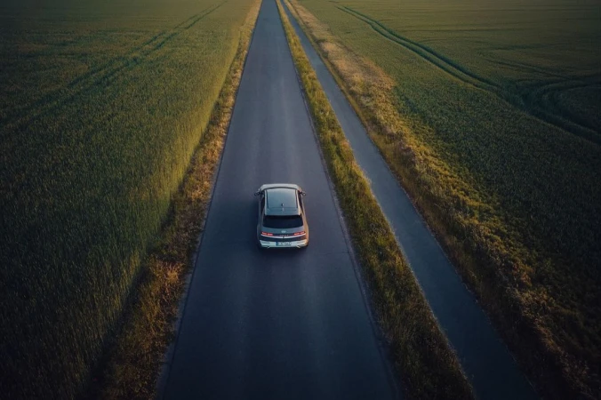 Overhead aerial shot of a gray car driving on a rural highway through green fields under soft, diffused light at dusk or dawn.