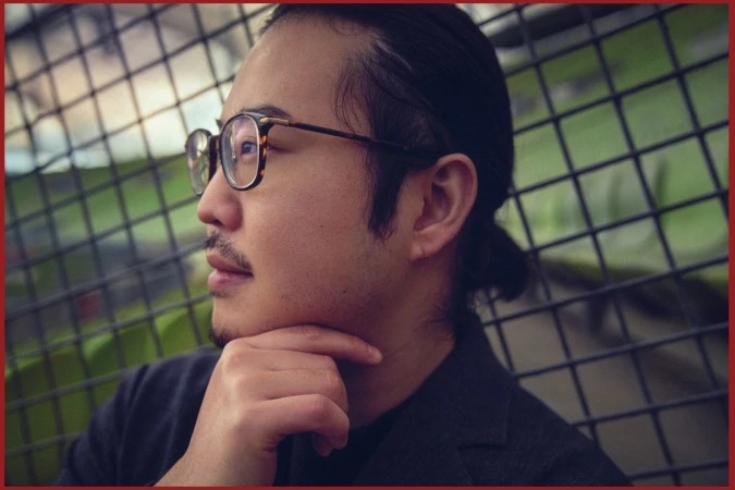Profile portrait of an East Asian man wearing tortoiseshell glasses and resting his chin on his hand against a metal grid fence