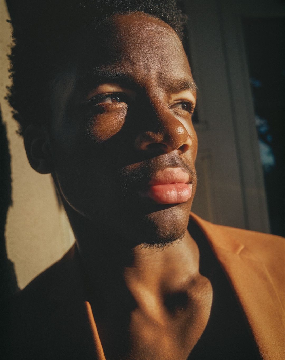Portrait of a young African American man in a tan blazer, gazing off into the distance with dramatic sunlight and shadow.