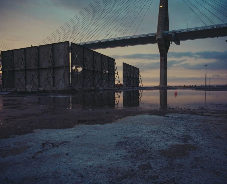 Low-angle shot of a large bridge pier under a cloudy sky at dusk. A puddle reflects the scene. In front is a barrier.