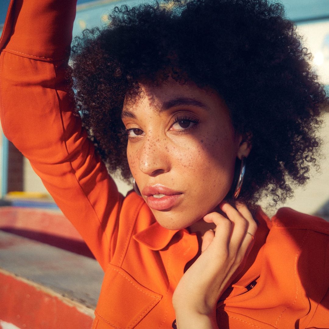 Close-up shot of a young woman with curly hair and freckles wearing an orange jacket, posing outdoors with a confident gaze.