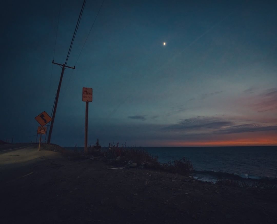 Evening landscape with ocean, utility poles along a dark road, and a vibrant sunset-streaked sky. Moody scene with coastal atmosphere.