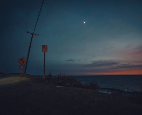 Evening landscape with ocean, utility poles along a dark road, and a vibrant sunset-streaked sky. Moody scene with coastal atmosphere.