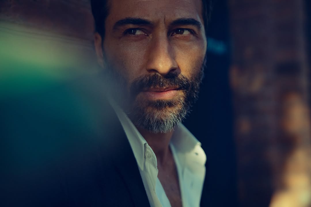 Close-up portrait of a middle-aged adult man with dark hair, a salt-and-pepper beard, an open white shirt, and a dark jacket.