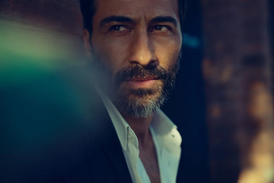 Close-up portrait of a middle-aged adult man with dark hair, a salt-and-pepper beard, an open white shirt, and a dark jacket.