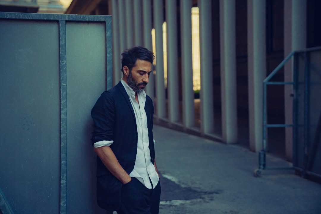 Adult man with beard in a navy blazer stands pensively in an urban setting next to a metal wall, looking down with hands in pockets.