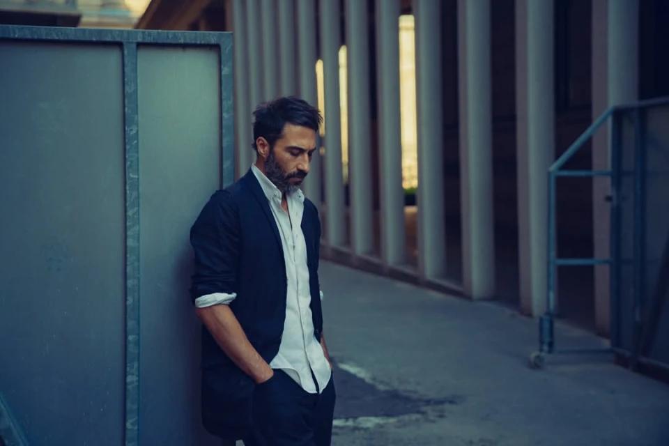 Adult man with beard in a navy blazer stands pensively in an urban setting next to a metal wall, looking down with hands in pockets.