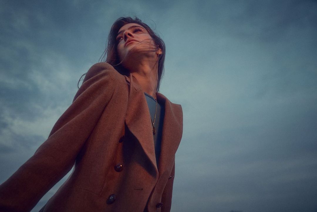 Portrait of a brunette woman in a brown double-breasted coat looking up toward the sky. She is outside against a cloudy blue sky.