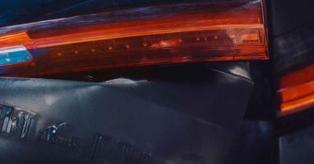 Automotive macro shot: a modern car's rear taillight and dark grey panel, illuminated by red-orange light, emphasizing detail and design.