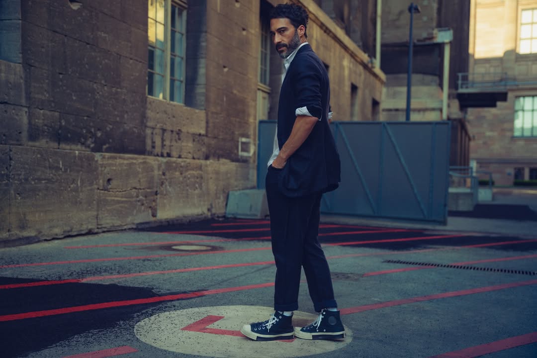 Man in a black suit stands outside in an urban setting with a serious expression, wearing high-top sneakers, against a brick building backdrop.