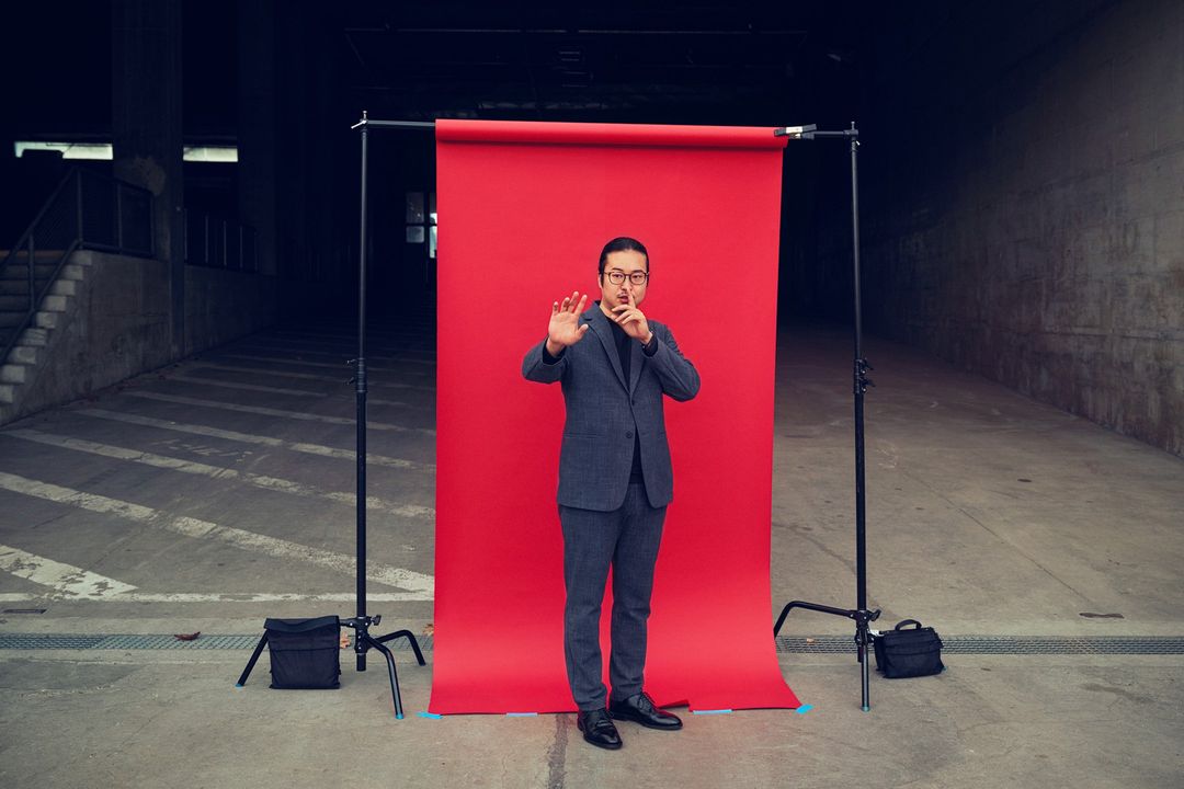Man in suit poses in front of a red backdrop set up outdoors. He wears glasses and holds his hand up with his fingers spread.