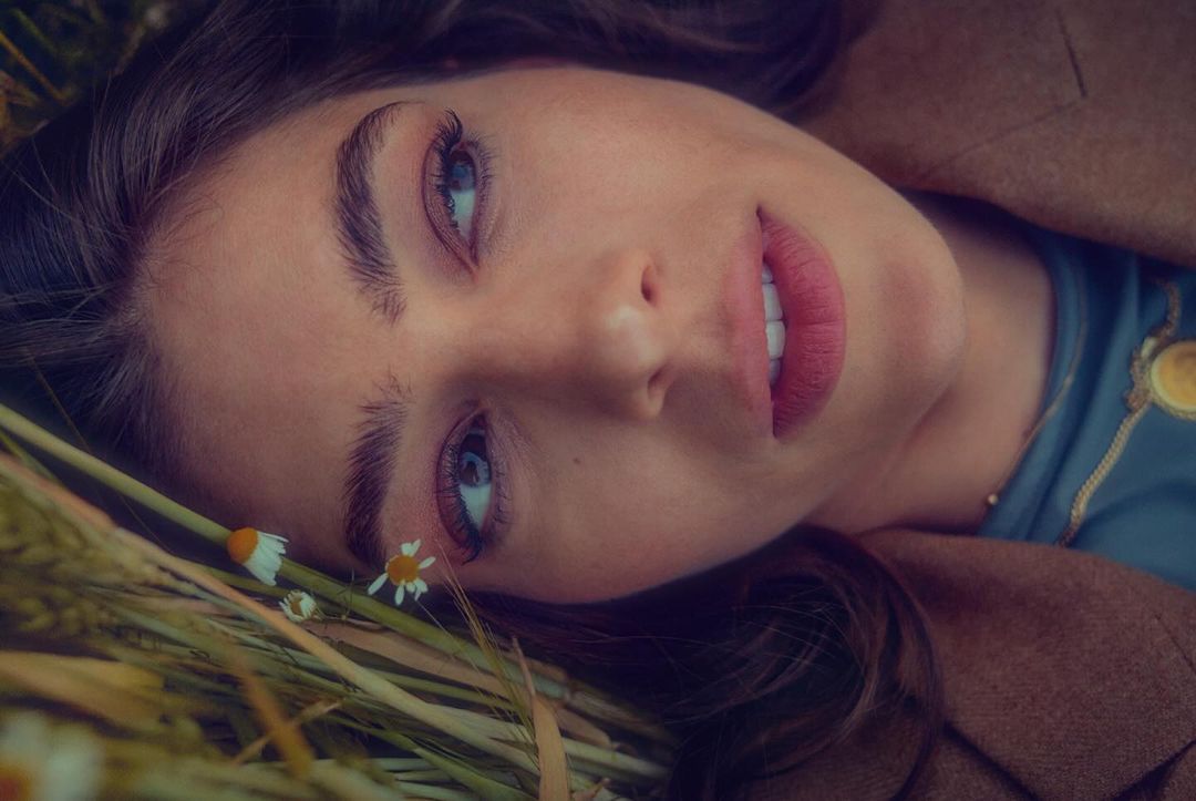 Close up portrait of a woman laying in a field of daisies wearing a brown jacket and teal shirt, smiling with soft lighting.