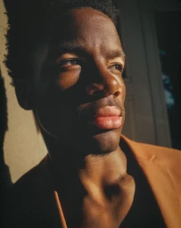 Close-up portrait of a young Black man in a camel jacket lit by warm sunlight casting sharp shadows across his face.