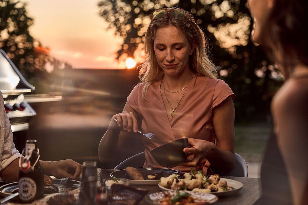 Outdoor dining scene at sunset featuring a blonde adult woman eating with friends, food on the table, and a barbeque in the background.