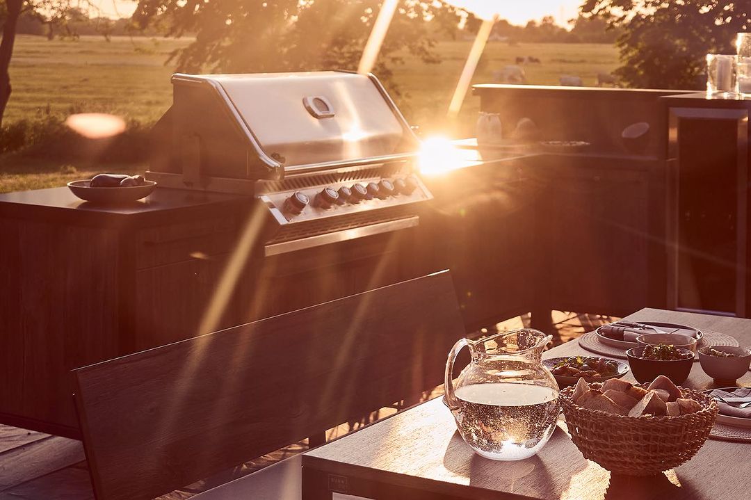 Outdoor dining table set with a pitcher of water and various dishes, a grill station and sunlight in a rural setting.