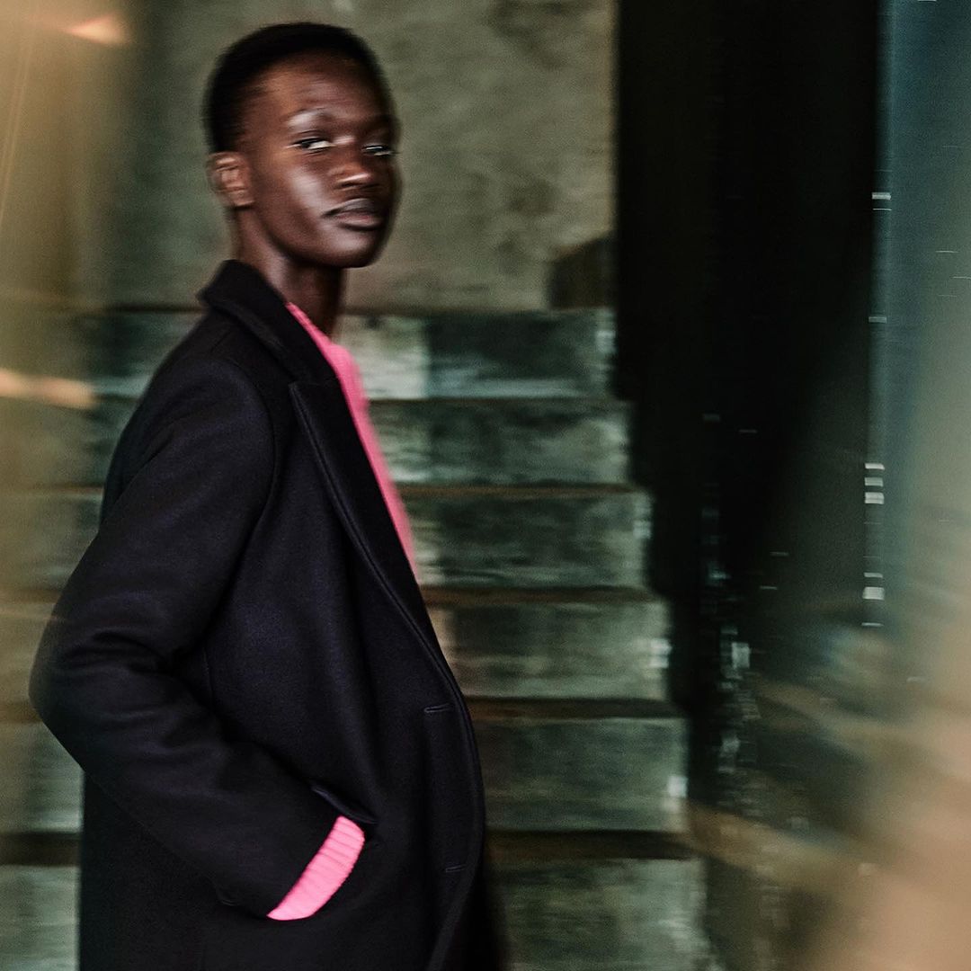 Stylish African-American man in a black coat and pink sweater stands indoors by stairs, looking over his shoulder.