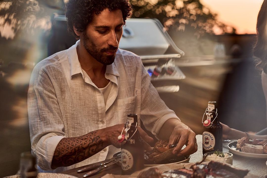 A man in a white shirt prepares food at a table with beer bottles, with a grill visible in the background, enjoying an outdoor meal.