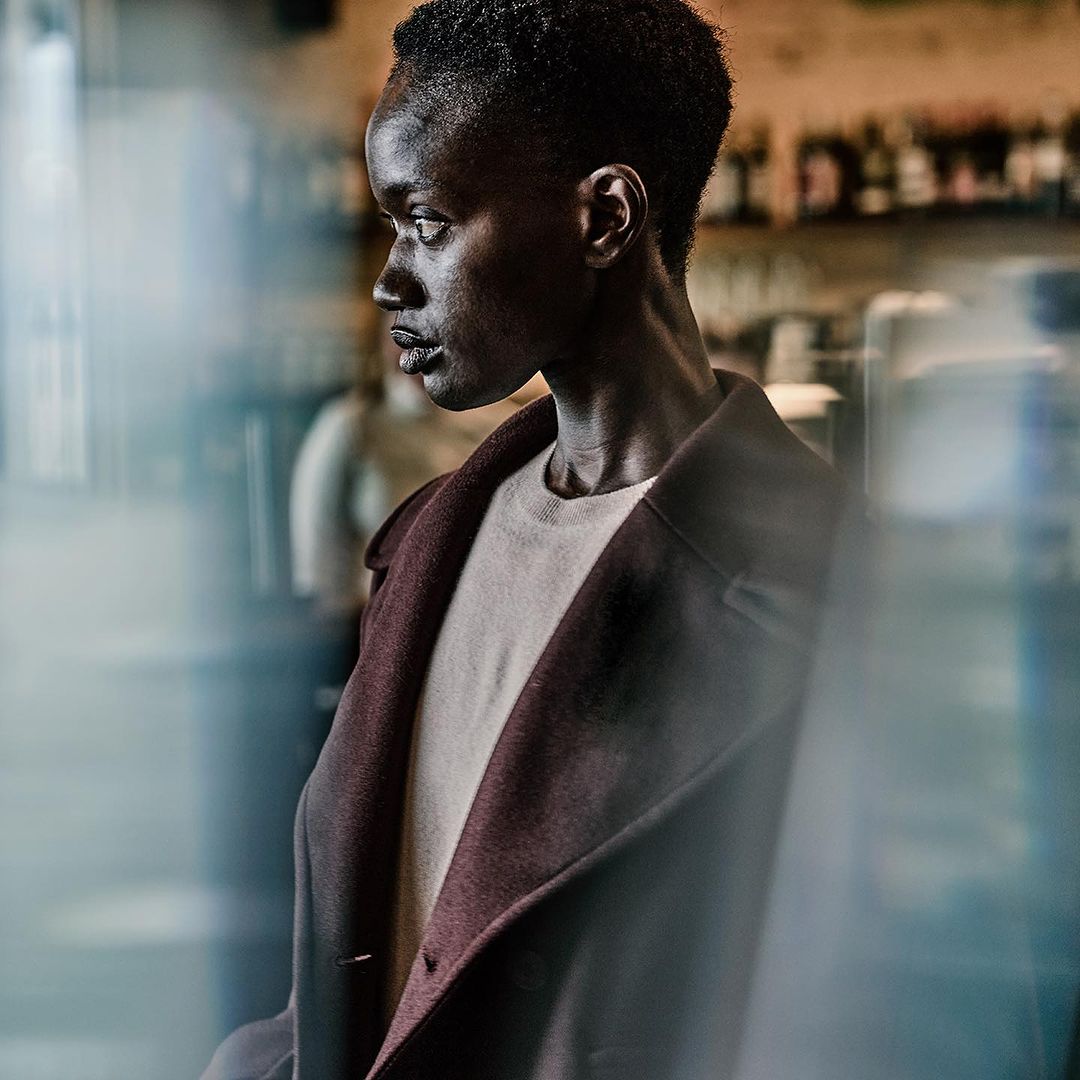 Portrait of an African woman in a coat looking left, with blurred interior background, casting a serious mood. Fashion editorial.