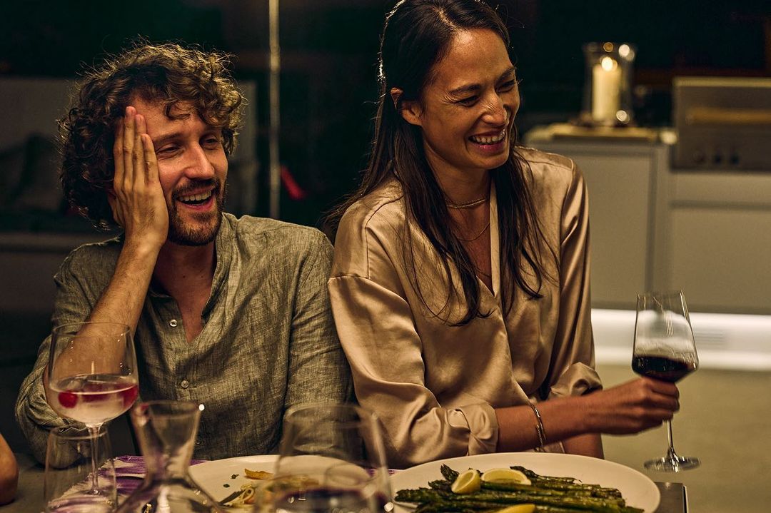 A happy couple laughs together at an indoor dinner party. They are sitting at a table with wine glasses and a plate of asparagus.