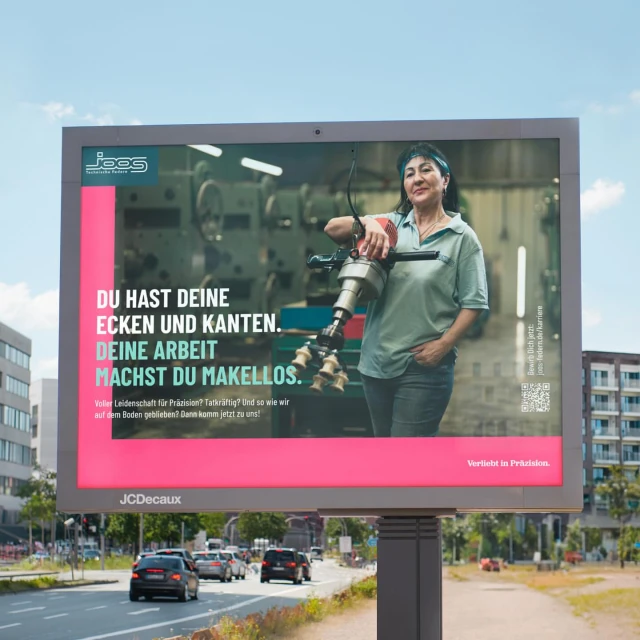 Billboard advertisement featuring a woman with an industrial tool in front of factory equipment and a pink text block against a bright sky.