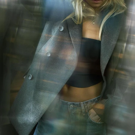 Close-up of woman wearing a grey pinstripe blazer, black bandeau top, and denim jeans, shot through glass reflection.