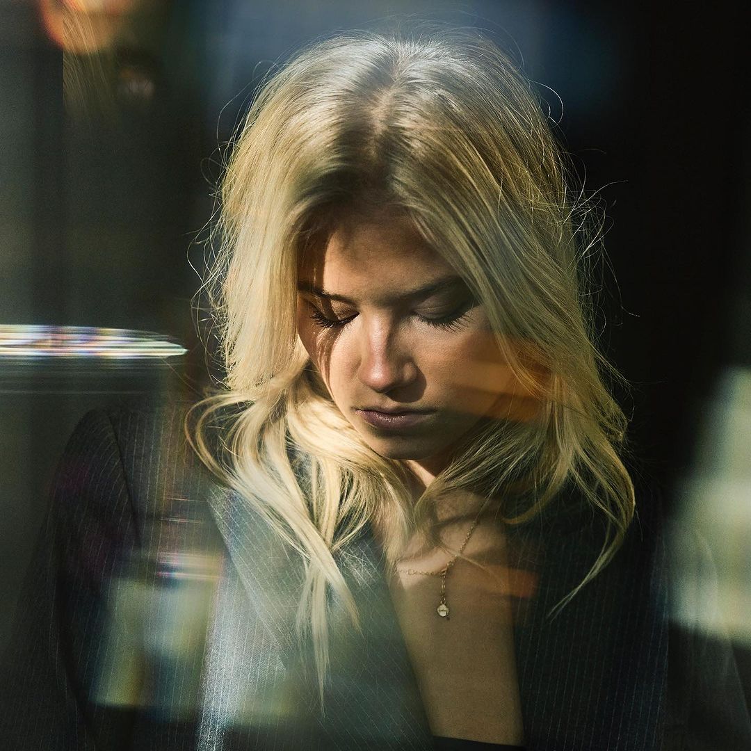 Close-up portrait of a blonde woman with her head tilted down, wearing a dark blazer and gold necklace, against a dark background.
