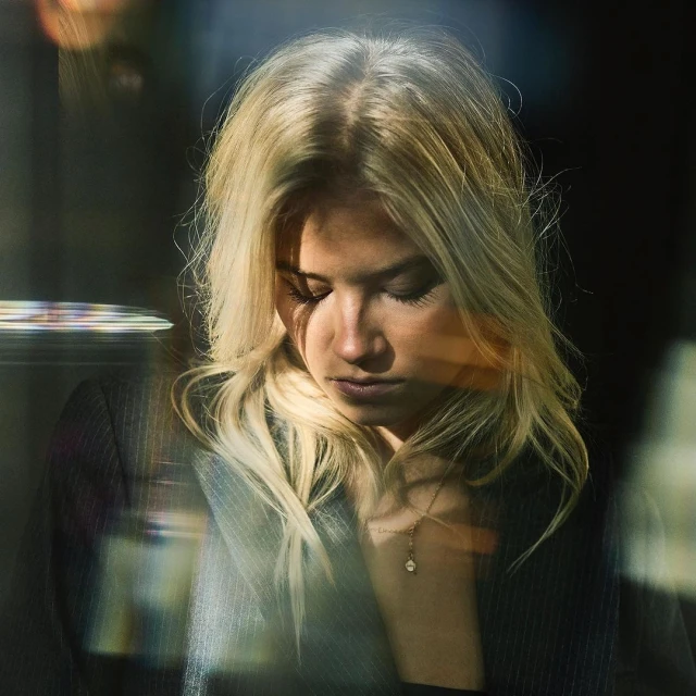Close-up portrait of a blonde woman with her head tilted down, wearing a dark blazer and gold necklace, against a dark background.