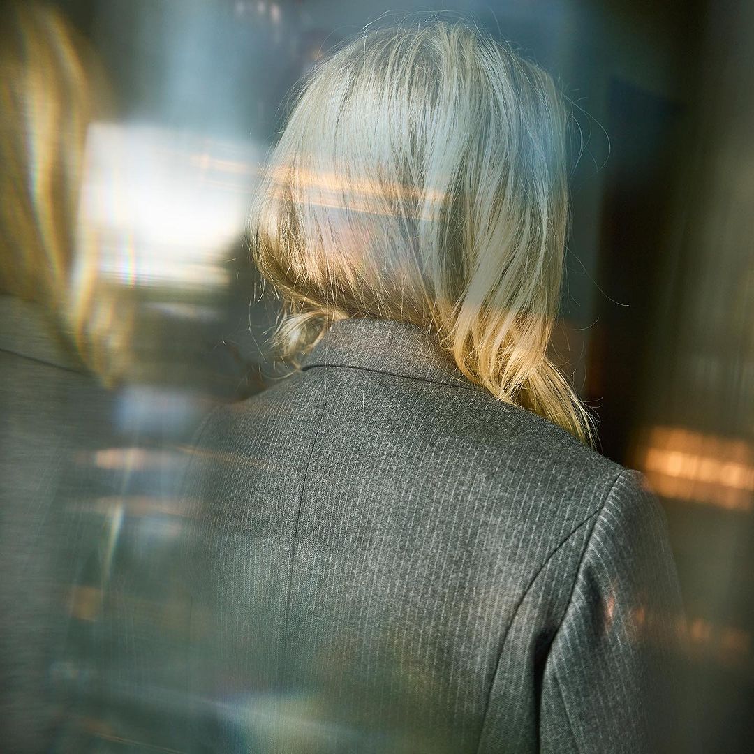 Back of woman with blonde hair wearing a grey pinstripe blazer in a blurry, abstract setting with refracted light and cool tones.