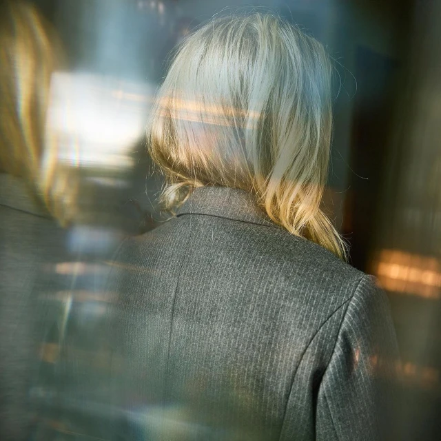 Back of woman with blonde hair wearing a grey pinstripe blazer in a blurry, abstract setting with refracted light and cool tones.