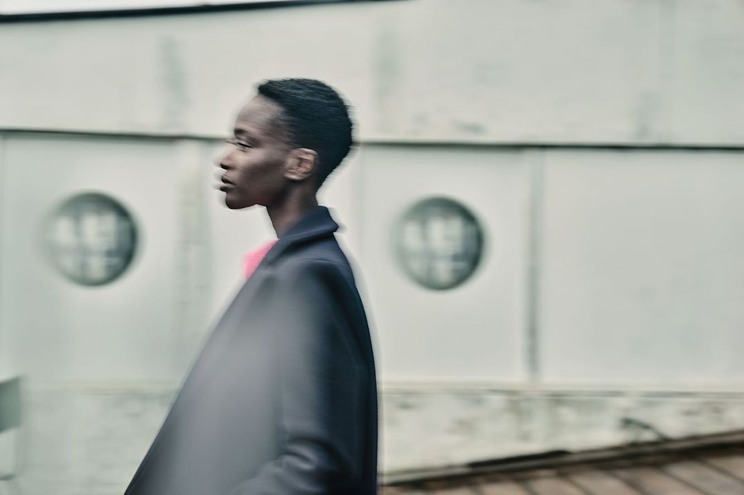 A blurry, medium shot of a Black woman in a dark coat, captured outdoors against a light wall with round window-like elements.
