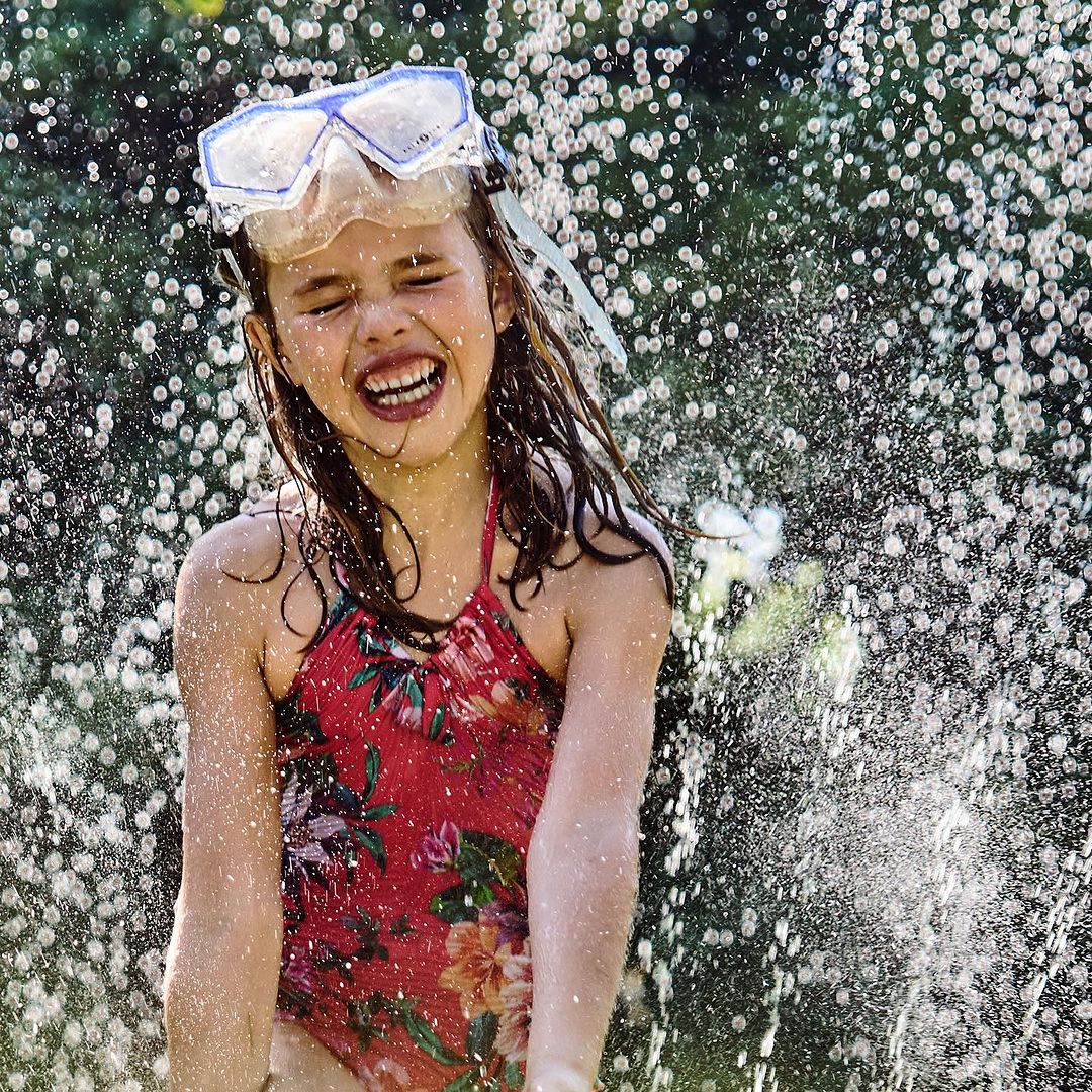 Young girl with brown hair and a floral bathing suit smiles while playing in water droplets with goggles on her head outdoors.