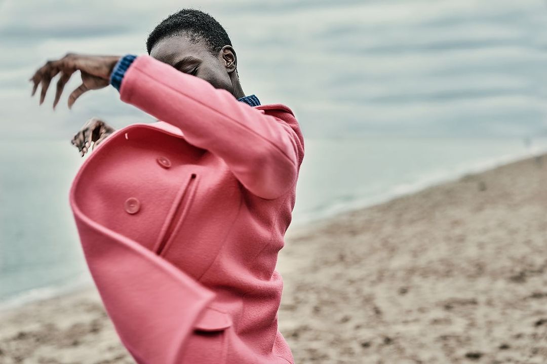 African-American man in pink coat poses expressively on a beach with ocean and cloudy sky in the background during daytime.