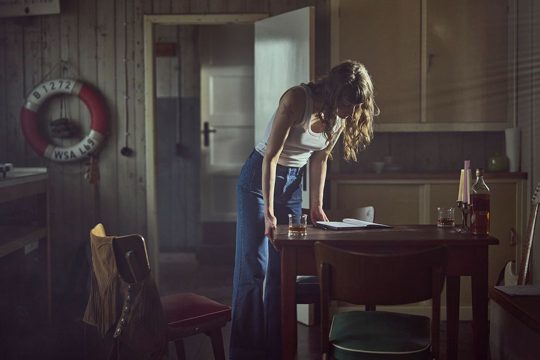 Woman stands at table with drink and notebook in dim, rustic interior. Life preserver visible on wood-paneled wall.