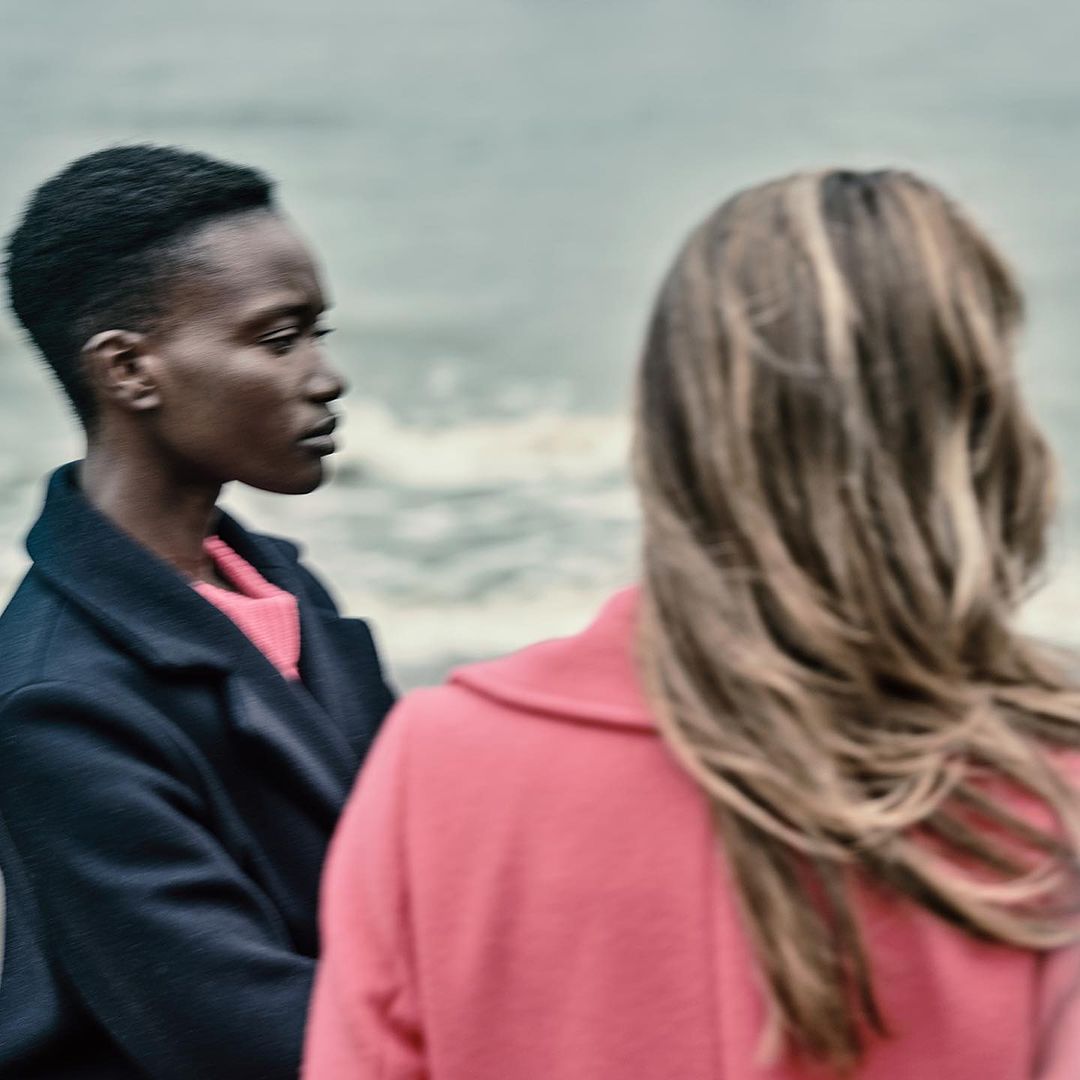 Two stylish women stand near a lake. One woman faces left, wearing a black coat. The other has long hair and wears a coral coat.