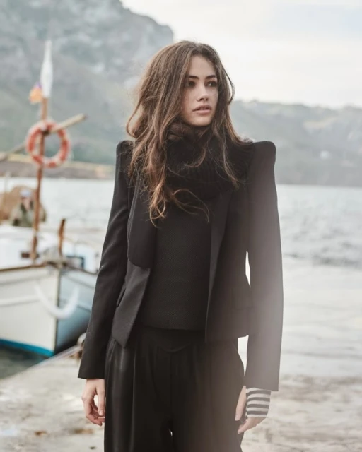 A young woman in a black suit poses on a concrete pier with a fishing boat and a distant, misty mountain range visible in the background.