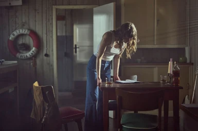 Woman in a white tank top and blue jeans leaning over a wooden table inside an interior room captured by Michael Heinsen.